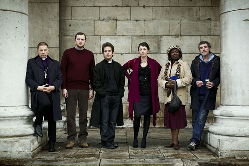 The cast of Rev outside the church, from left to right: Simon McBurney as the Archdeacon, Miles Jupp as lay reader Nigel, Tom Hollander as Reverend Adam Smallbone, Olivia Colman as Alex Smallbone, Ellen Thomas as Adoha, and Steve Evets as Colin
