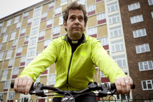 Tom Hollander as the Reverend Adam Smallbone on his bike, wearing a yellow luminous jacket