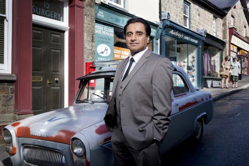 Dr Prem Sharma (Sanjeev Bhaskar) stands beside a car outside the shops in Trefelin