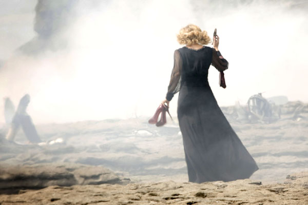 River Song, played by Alex Kingston, walks across the beach with red high heels in hand
