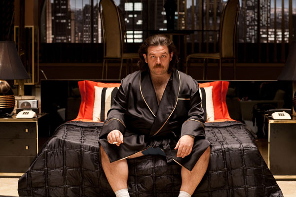 Nick Frost as John Self sits in his hotel room